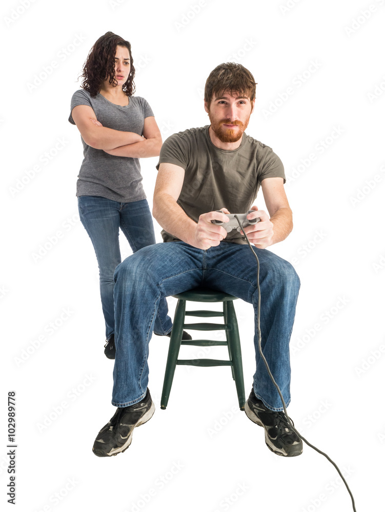 Photograph of a young man in a tee shirt absorbed in a video game. His girlfriend watches him from behind with a pained look. Photograph of a young man in a tee shirt absorbed in a video game. His girlfriend watches him from behind with a pained look.