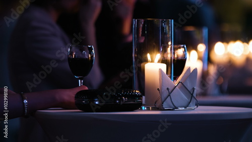 Close-up footage of elegantly decorated table at restaurant, woman holding a glass of red wine, fancy black clutch bag is near. Man brings a plate with food and have a drink.  