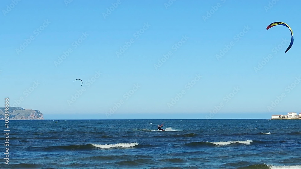 Kite surfers glide over the blue waters of Pollenca Bay, Majorca Island. Late afternoon, the low sun lights up the resort on the other side of the bay Kite surfers glide over the blue waters of Pollenca Bay, Majorca Island. Late afternoon, the low sun lights up the resort on the other side of the bay
