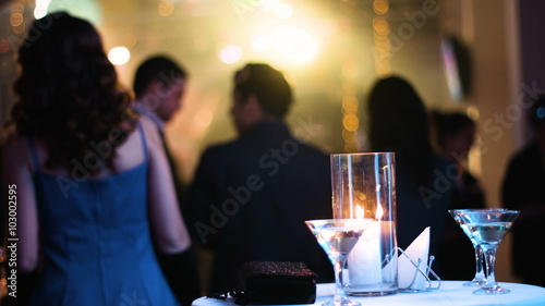 Table with a couple of cocktail glasses on it, candle is burning and black clutch bag lying aside, people standing back to camera and watching a concert on a stage at nightclub 