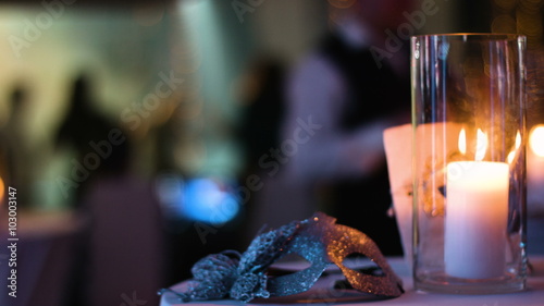Masquerade gold venetian mask lies on a table in a nightclub during a costume party, white candle burning in a glass cup,  blurred footage of people partying on a background.