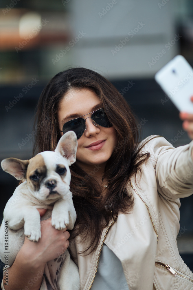 Beautiful young girl smiling and taking a selfie with her cute French bulldog puppy. Urban scene. Selective focus on girl. Beautiful young girl smiling and taking a selfie with her cute French bulldog puppy. Urban scene. Selective focus on girl.