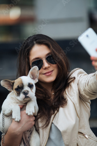 Beautiful young girl smiling and taking a selfie with her cute French bulldog puppy. Urban scene. Selective focus on girl. photo
