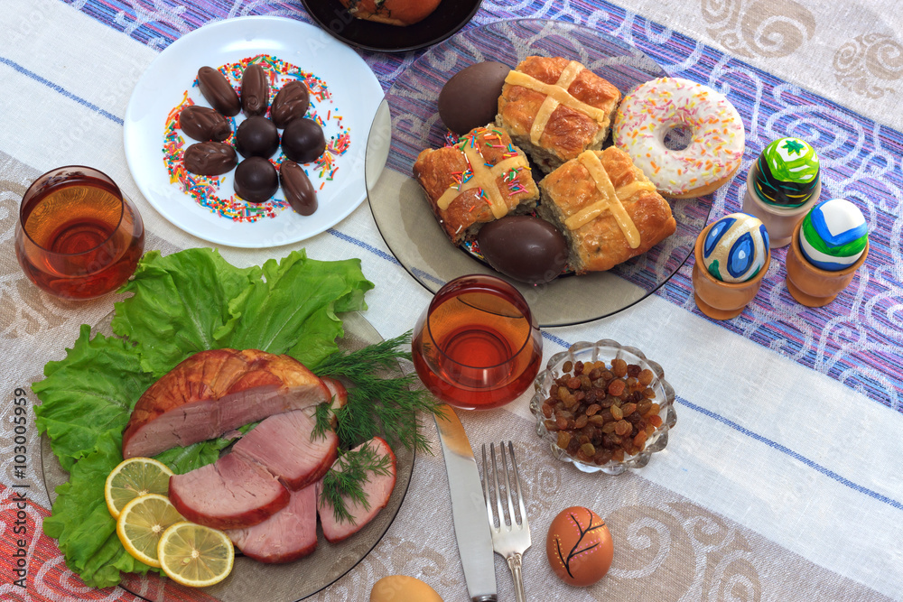 Traditional easter dinner set with sliced meat with lemon and herbs, handmade colored eggs, chocolates, raisins, easter cake and glasses of juice on colorful tablecloth Traditional easter dinner set with sliced meat with lemon and herbs, handmade colored eggs, chocolates, raisins, easter cake and glasses of juice on colorful tablecloth