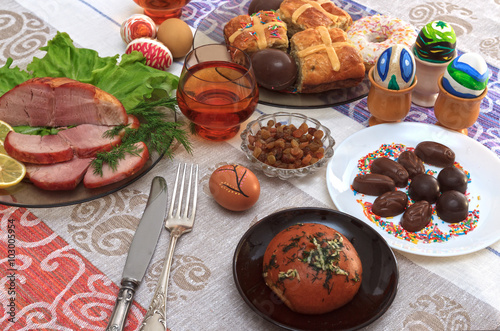 Traditional easter dinner set with sliced meat with lemon and herbs, bread, handmade colored eggs, chocolates, raisins, easter cake and glasses of juice on colorful tablecloth