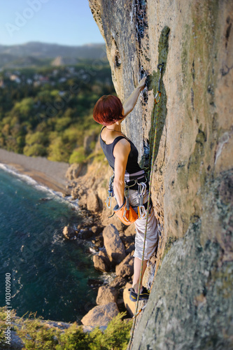 Athletic girl climbing an overhanging cliff against the scenic sea coast background. Holding one hand in magnesium bag. Summer time.