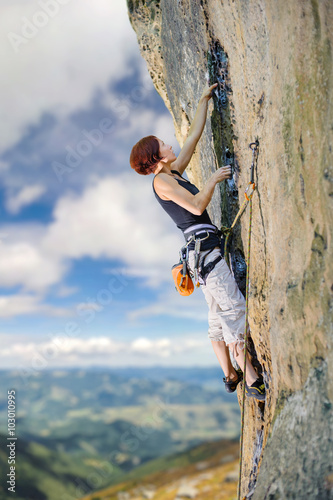 Young female rock climber climbing with rope and carbines an overhanging cliff against blue sky and mountains background. Summer time.