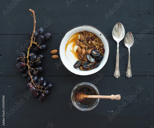 Healthy breakfast set. Bowls of oat granola with yogurt, fresh grapes, almond and honey over black wooden backdrop