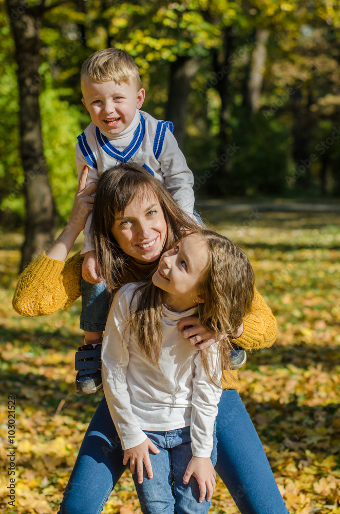 Smiling mother hugs daughter and holds son on her shoulders in the autumn park Smiling mother hugs daughter and holds son on her shoulders in the autumn park