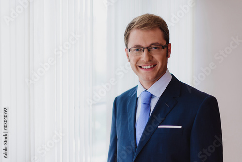 Portrait of a smiling businessman, dark blue suit with a blue tie and glasses, standing in office in front of windows.