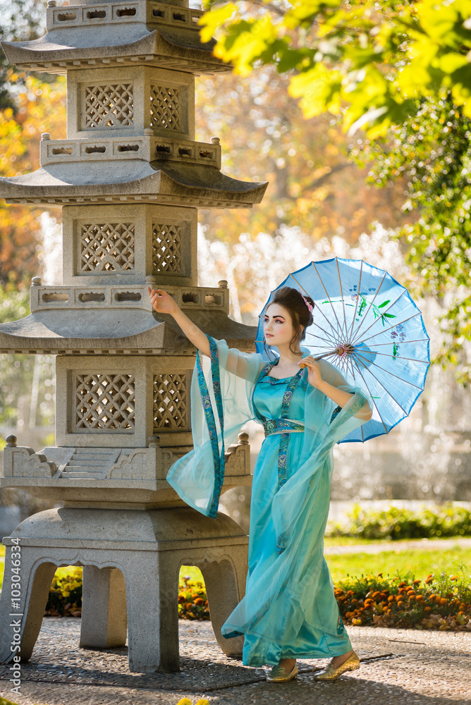 Fototapeta premium beautiful geisha with a blue umbrella near stone pagoda on background of blur fountain