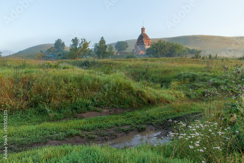 Summer landscape with dirt road and destroyed the old rural church on the hilly bank of the river a sunny misty morning
