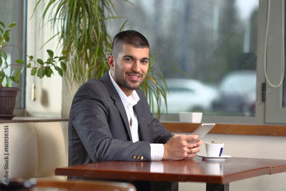 Smiling young businessman using his digital tablet while having a cup of coffee in a restaurant Smiling young businessman using his digital tablet while having a cup of coffee in a restaurant