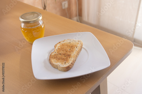 Tostada y tarro de miel en un plato blanco con luz natural encima de una mesa. Desayuno.