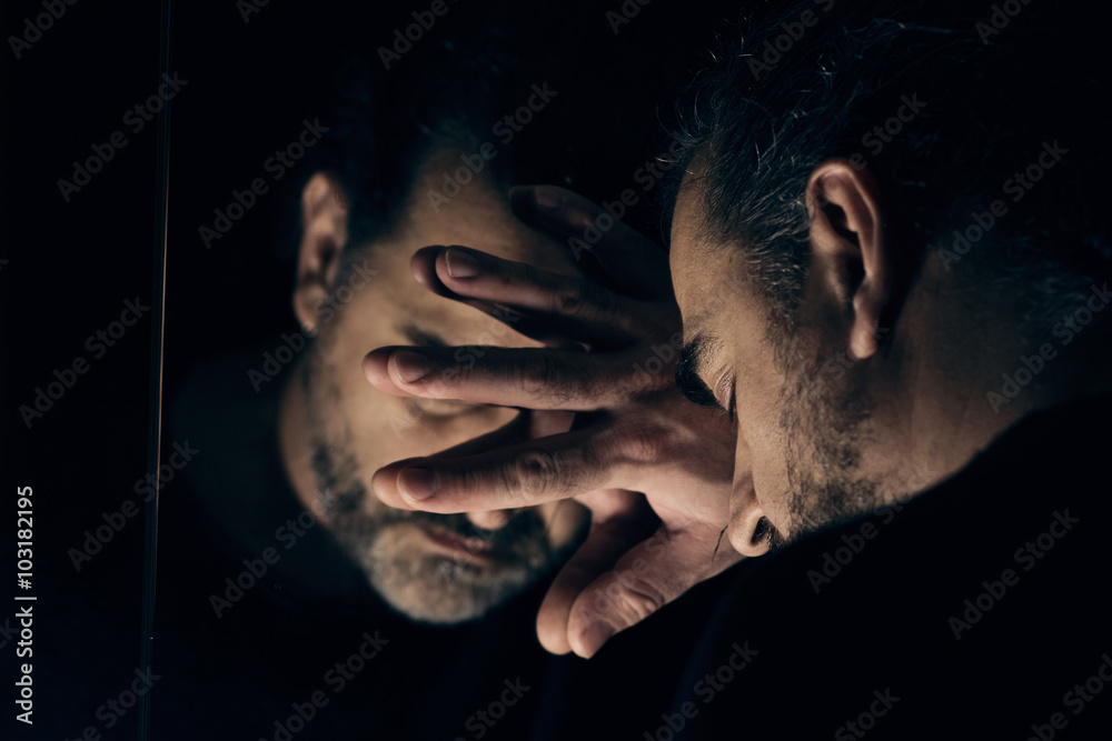 Man in despair, sad and lonely resting his head on a hand, in front of mirror