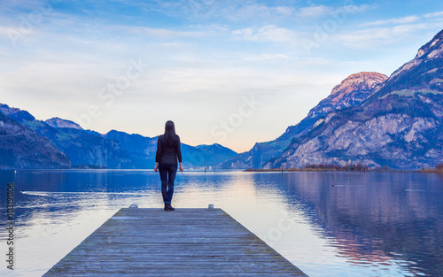 Epic mountain landscape. Female figure on a wooden pier. Lake at sunset.