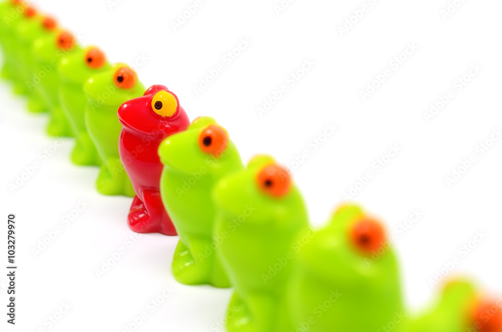 Obraz premium Line of green toy frogs. Among them, one red frog. Close up view. Different from the others. At home among strangers. Concept of antagonism.