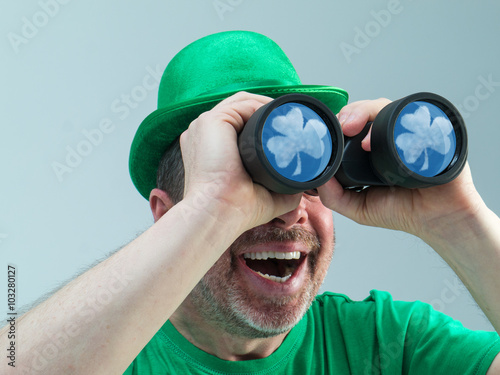 Photograph of a St. Patrick's Day reveler looking into the sky with binoculars. Reflected in the lenses are a shamrock shaped cloud. photo