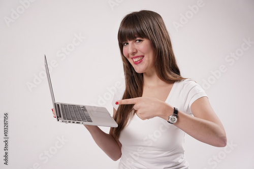 Eine junge Frau, Mädchen sitzt barfuß auf dem Boden und hält einen Laptop in einer Hand. Mit der anderen zeigt sie mit einem Finger auf den Bildschirm