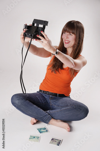 Eine junge Frau / Mädchen sitzt barfuß im Schneidersitz auf einem Boden und macht mit einer Sofortbildkamera - Polaroidkamera ein Selfie - Selbstportrait. Vor ihr liegen weitere Polaroids 
