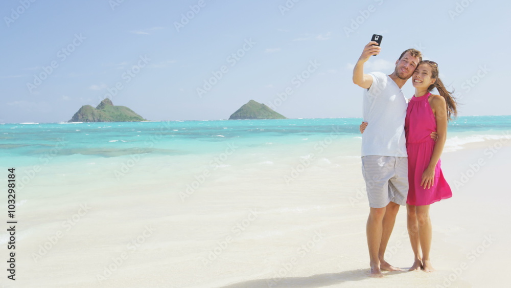 Couple on beach vacation taking selfie photograph using smart phone. Romantic couple in love on honeymoon on Lanikai beach, Oahu, Hawaii, USA with Mokulua Islands. Couple holding smartphone camera. Couple on beach vacation taking selfie photograph using smart phone. Romantic couple in love on honeymoon on Lanikai beach, Oahu, Hawaii, USA with Mokulua Islands. Couple holding smartphone camera.