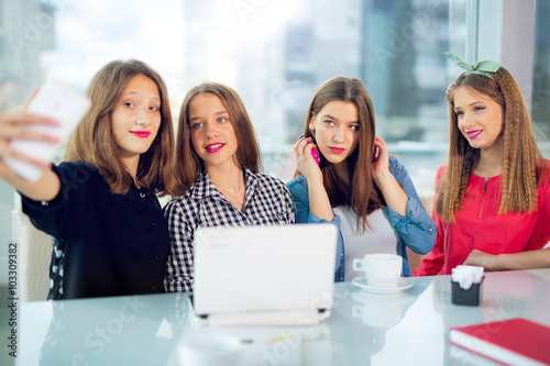 Portrait of four young women sitting at the table in the cafe take selfie with smart phone