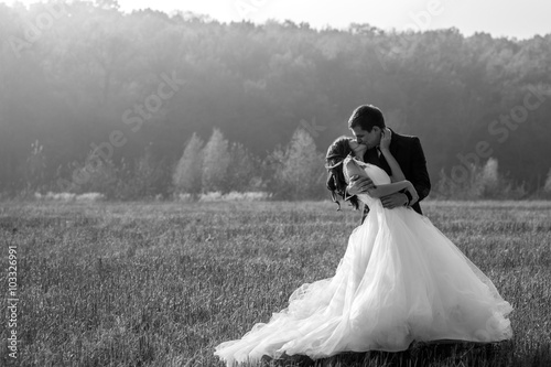 Romantic fairytale newlywed couple hug & kiss in field at sunset