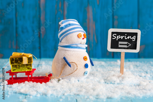 Spring change winter background. Snowman with gift and red sled stand near direction sign spring. Postcard photo