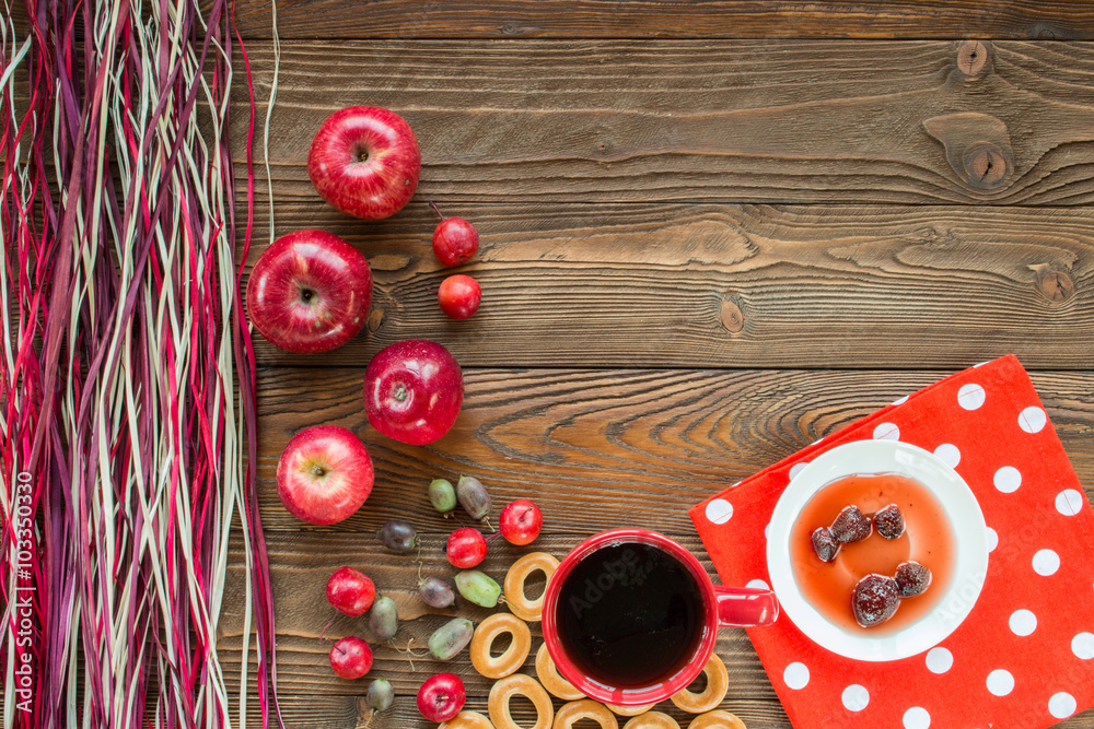 red cup of tea, ripe red apples, actinidia berries, bagels, strawberry jam in white saucer, spoon, red napkin at white polka dots on wooden table with decorative colorful  dry straw. top view