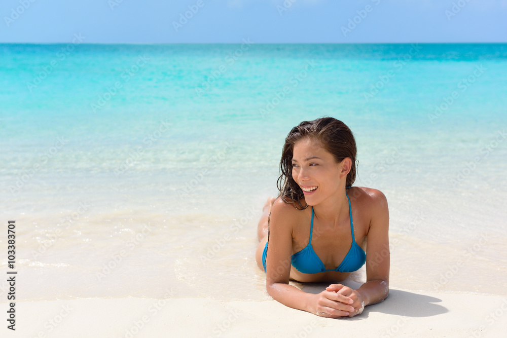 Beach vacation woman relaxing on sand happy. Asian mixed race model looking happy lying down on perfect white sand beach and turquoise ocean water for luxury summer vacations in exotic destination.