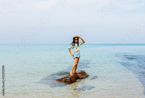 Attractive Asian woman standing on rock on sandy beach, smiling and enjoying a beautiful sunny day