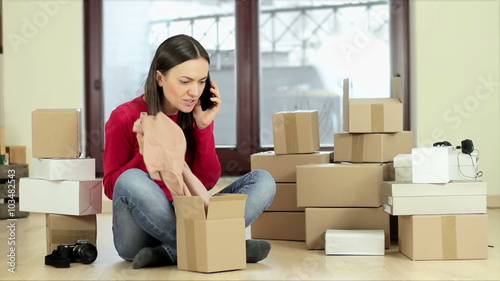 Young woman looks disappointed after shopping on line and complaining by phone with laptop and stack of boxes on the floor