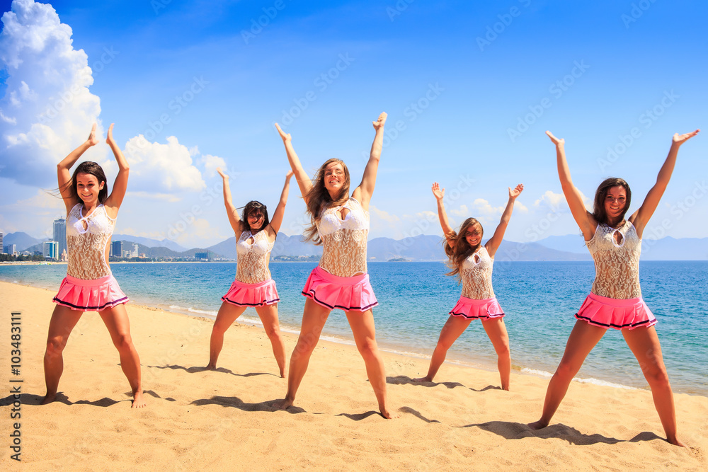 cheerleaders dance on beach with hands upwards against azure sea