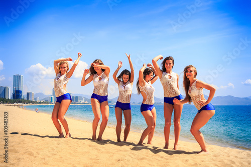 cheerleaders stand on beach laugh wave hands against azure sea