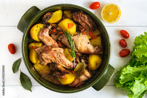 Top flat view of roasted rabbit meat with vegetables in round ceramic pot on white wooden table surface. Food ingridients