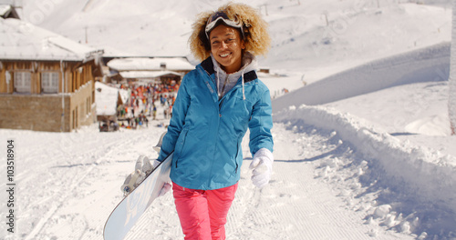 Smiling pretty young woman with a trendy afro hairstyle carrying a snowboard standing in fresh winter snow at a mountain ski resort