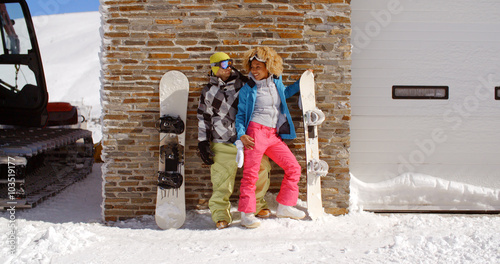 Cute couple in snowsuits standing next to upright snowboards while they lean against ski resort garage with tractor in thick snow on sunny day