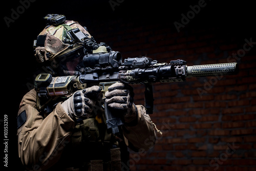 Spec ops soldier in uniform with assault rifle/man in military uniform with assault rifle aiming at target on background of dark wall