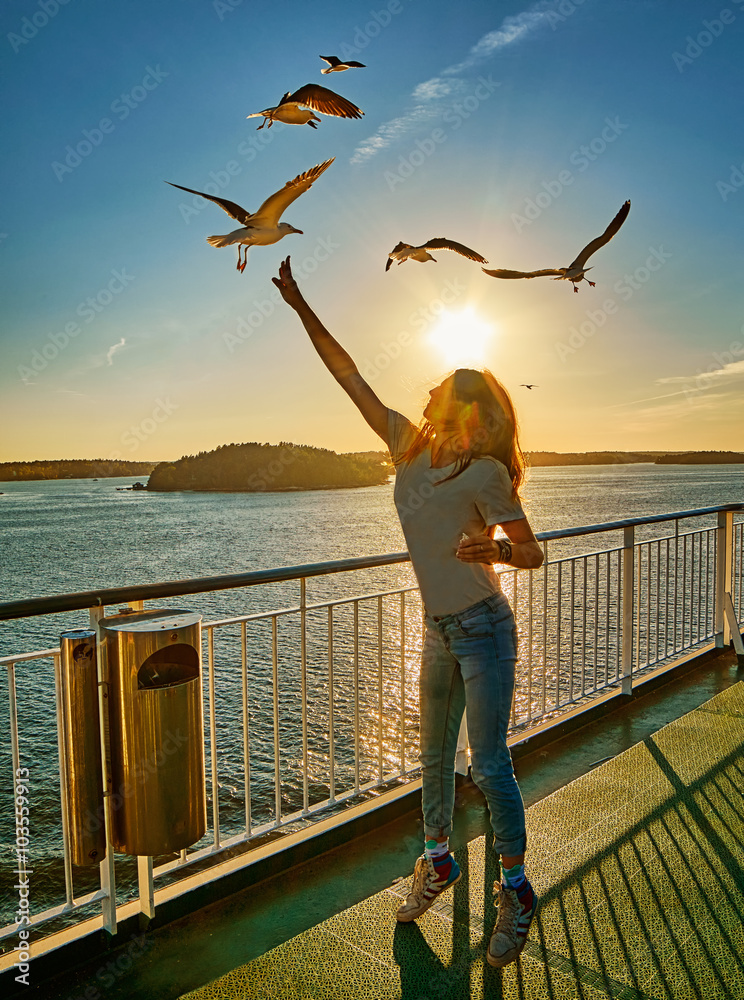 Fototapeta premium Young girl feeding seagulls in the flare of setting sun onboard a ferry in Scandinavia