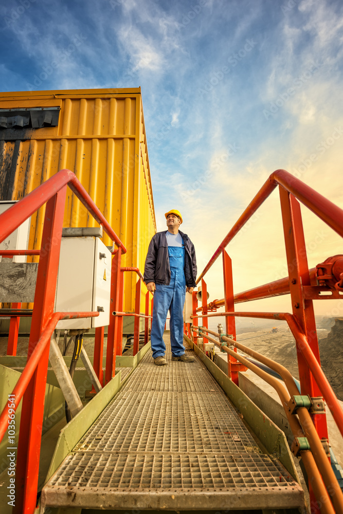 Smiling and confident engineer with helmet standing on huge drilling machine and looking at side. Mining site in background. Smiling and confident engineer with helmet standing on huge drilling machine and looking at side. Mining site in background.