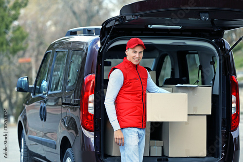 Young handsome delivery man standing near the car with boxes and packages, outdoors