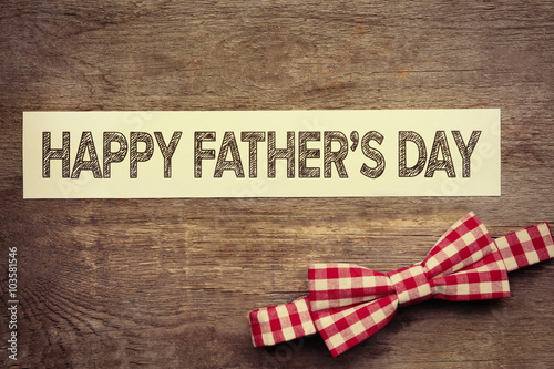 Happy Father's Day inscription with red bow tie on wooden background. Greetings and presents
