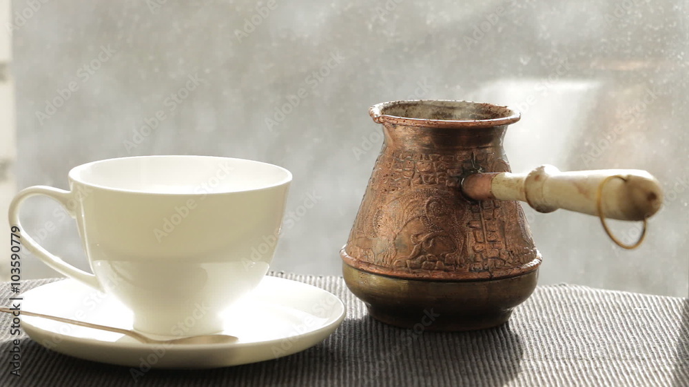 A brass jezve and a porcelain cup are in front of a window. Winter with snow trees outside. Cup and jezve are reflected in the window. Hot coffee is steaming. A brass jezve and a porcelain cup are in front of a window. Winter with snow trees outside. Cup and jezve are reflected in the window. Hot coffee is steaming.