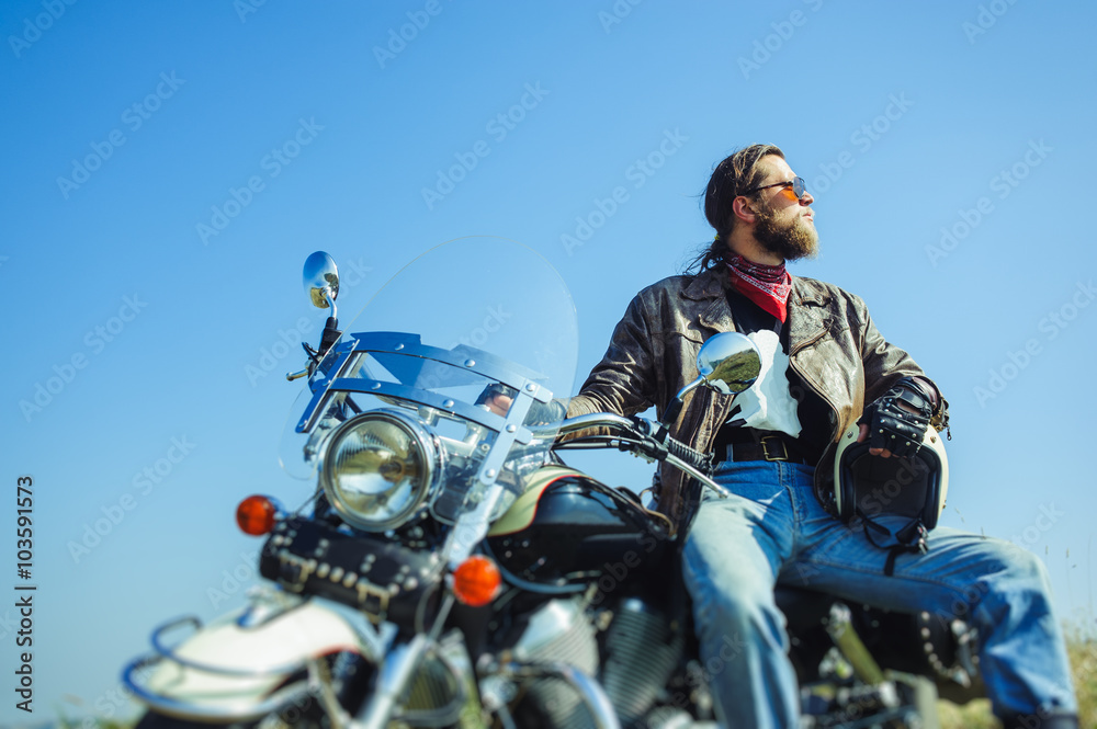 Portrait of a young man with beard sitting on his cruiser motorcycle. Man is wearing leather jacket and blue jeans. Low point of view. Tilt shift lens blur effect Portrait of a young man with beard sitting on his cruiser motorcycle. Man is wearing leather jacket and blue jeans. Low point of view. Tilt shift lens blur effect