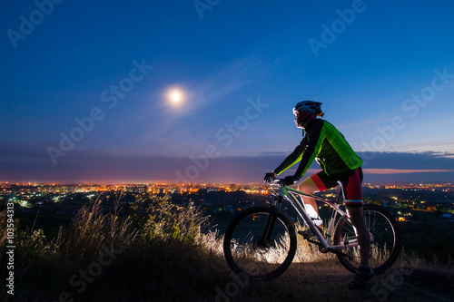 Young athlete biker with mountain bike on top of the hill observing the evening city view. Man is wearing green jacket
