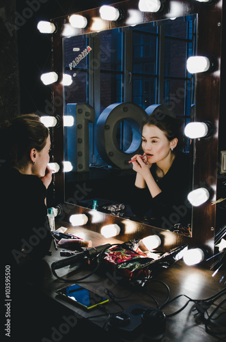 Young beautiful woman applying her make-up lips, looking in a mirror, sitting on chair at Theatre dressing room with vintage mirror with bulbs dark room