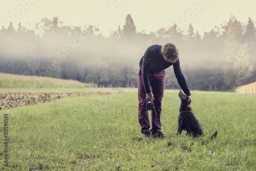 Fotografi Man standing in green grassland bending down to pat his black do