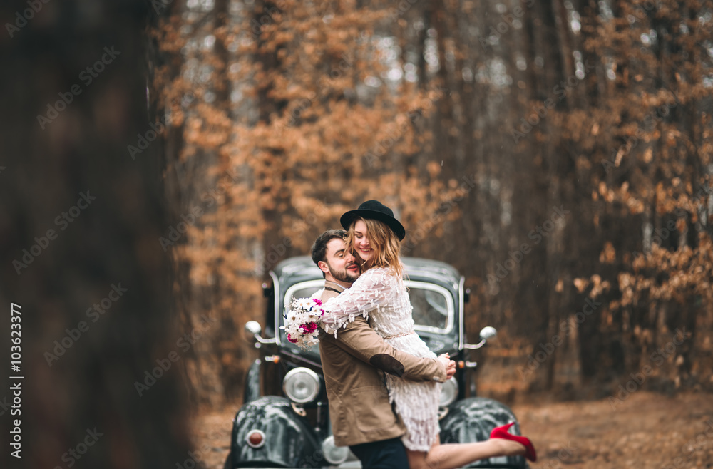 Fototapeta premium Stylish Loving wedding couple kissing and hugging in a pine forest near retro car
