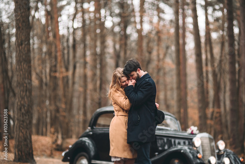 Stylish Loving wedding couple kissing and hugging in a pine forest near retro car
