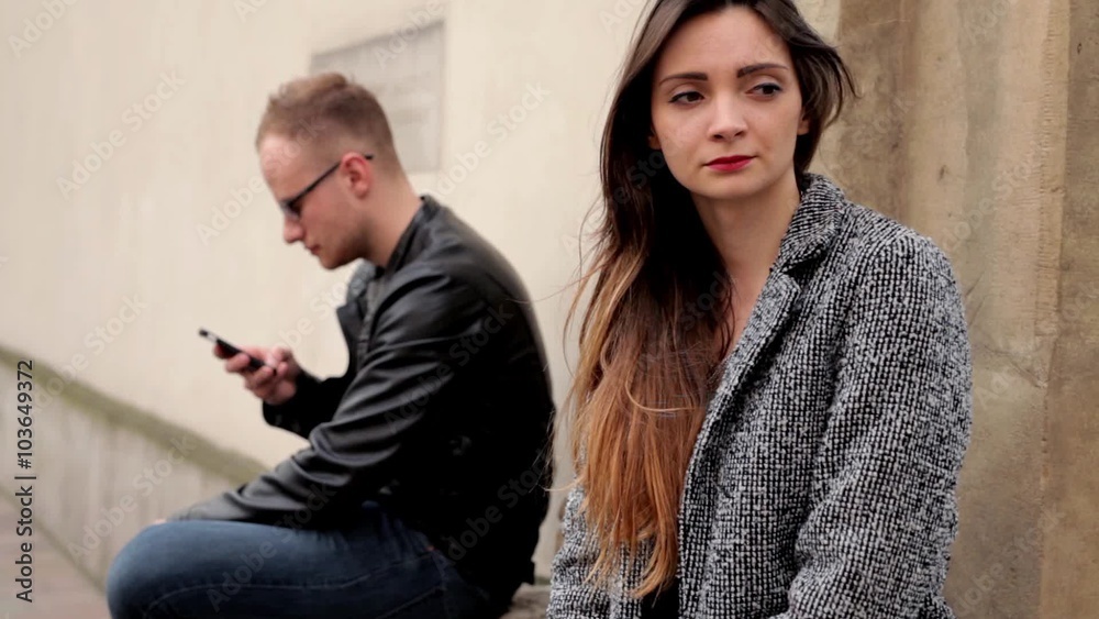 Young, brown hair girl sitting on the stone wall next to the boy in glasses. He is on the background and using phone. Girl is sad. Young, brown hair girl sitting on the stone wall next to the boy in glasses. He is on the background and using phone. Girl is sad.